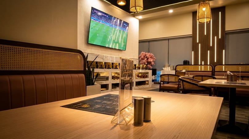 Restaurant interior with light wood tables, rattan-backed booths, and a large TV showing a soccer game.