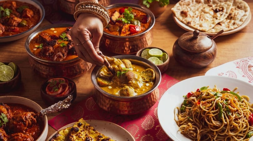 A hand with bangles dips food into a bowl of Indian curry amidst a spread of dishes.